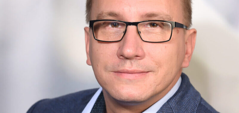 Ein Mann mit kurzen braunen Haaren und leichten Bartstoppeln, der ein blaues Hemd mit Kragen trägt, lächelt sanft in die Kamera vor einem neutralen grauen Hintergrund.