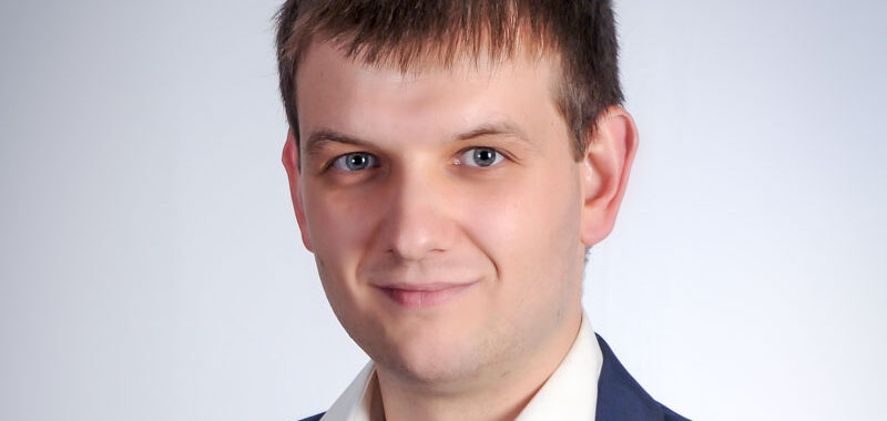 Ein Mann mit kurzen braunen Haaren, der eine dunkle Anzugsjacke und ein weißes Hemd trägt, lächelt vor einem schlichten hellgrauen Hintergrund.