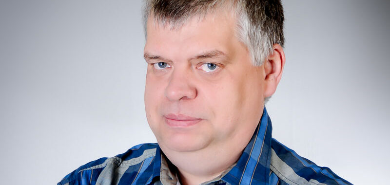 Ein Mann mittleren Alters mit kurzen grauen Haaren und blauen Augen, der ein blau kariertes Hemd trägt, posiert für ein Studioporträt vor einem schlichten hellen Hintergrund.