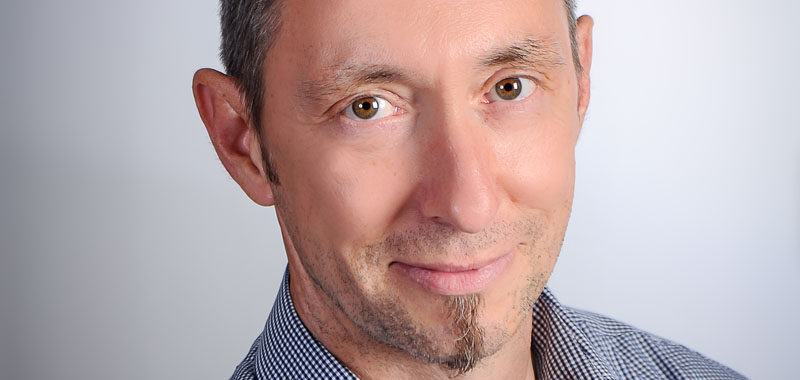 Ein Mann mit kurzen braunen Haaren und einem markanten Ziegenbart lächelt sanft in die Kamera. Er trägt ein kariertes Hemd und posiert vor einem schlichten hellen Hintergrund.