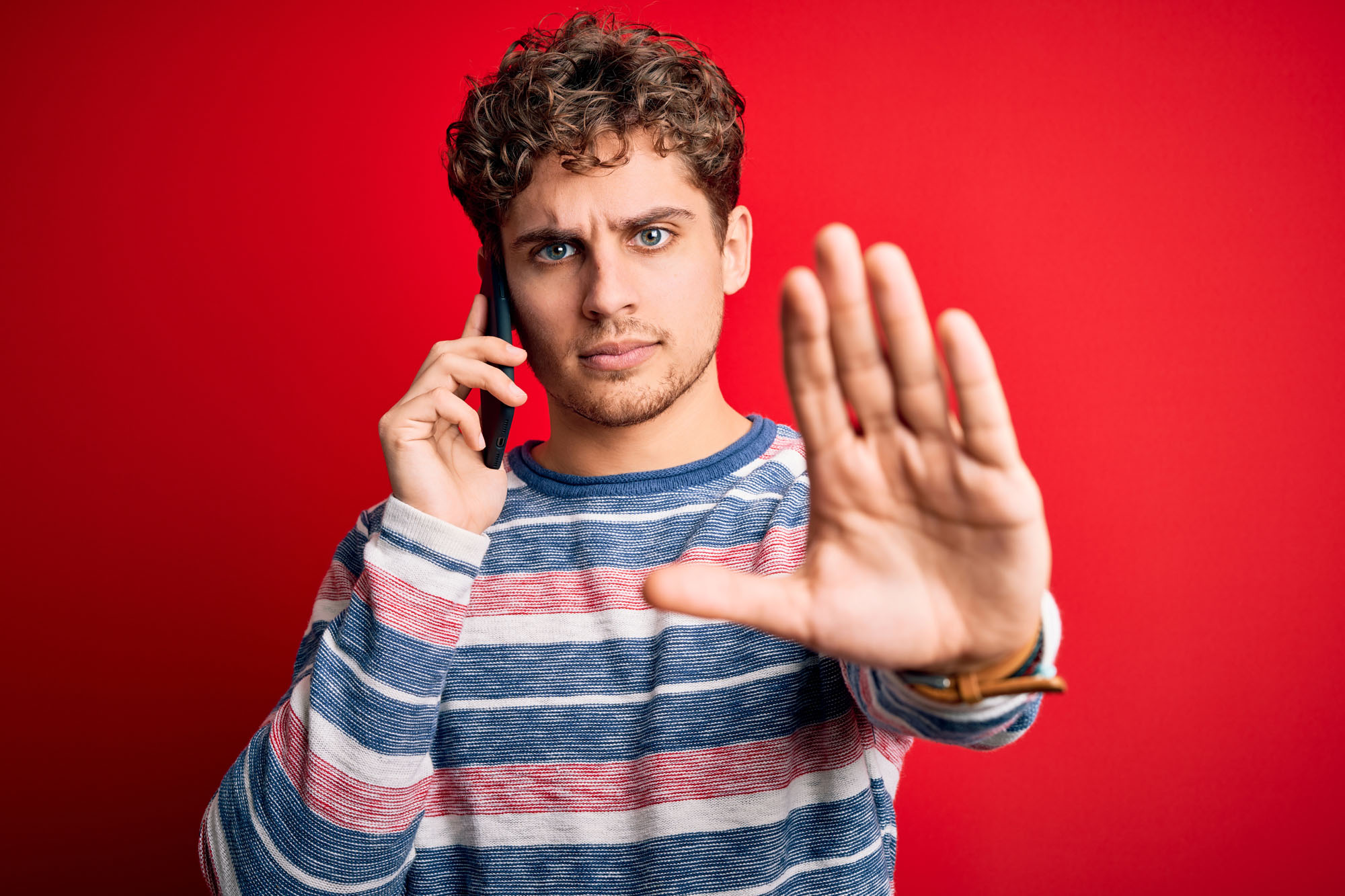 Young blond man with curly hair having conversation talking by the smartphone with open hand doing stop sign with serious and confident expression, defense gesture Ein junger Mann mit lockigem Haar und einem gestreiften Pullover hält sich ein Telefon ans Ohr und hebt die andere Hand in einer Stopp-Geste, während er vor einem leuchtend roten Hintergrund steht.